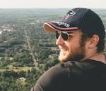 Smiling man in sunglasses and cap enjoying a scenic outdoor view from a hilltop.